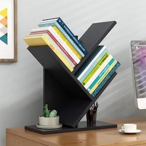 Op de afbeelding: Een zwart, boomvormig boekenrek gevuld met kleurrijke boeken. Het boekenrek heeft meerdere schuine planken en staat op een houten bureau. Een kleine potplant en een kopje koffie staan ook op het bureau.