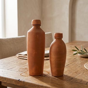 May include: Two terracotta bottles of varying sizes are displayed on a wooden table. The bottles have a cylindrical form with a rounded top. The table is made of wood and has a natural finish. The bottles are a warm, earthy tone.