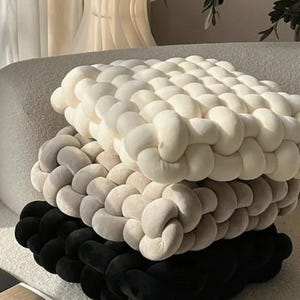 Puede incluir: Tres cojines cuadrados decorativos apilados. El cojín superior es blanquecino, el cojín central es beige y el cojín inferior es negro. Cada cojín está hecho de un material suave y anudado, creando una superficie texturizada.