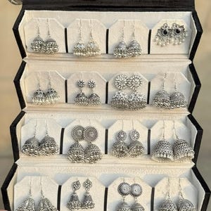 May include: A black jewelry case displaying various silver-toned earrings. The earrings feature diverse designs, including beaded, floral, and bell-shaped styles. The case is held open, showcasing multiple pairs of earrings.