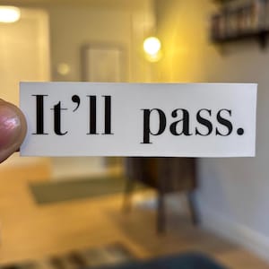 May include: A white rectangular sticker with the phrase "It'll pass." in black, serif font. The sticker is held up in front of a blurred interior background, suggesting a home or office setting.