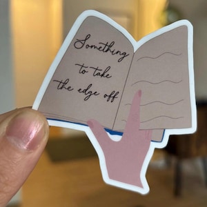 以下が含まれることがあります： 「Something to take the edge off」という黒い筆記体で書かれたテキストが特徴の、開いた本が描かれたステッカー。本はベージュ色のページと青い背表紙があり、手が本を持っています。