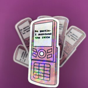 May include: A sticker featuring a pink flip phone design with the text "Be gentle - I survived the 1900s." The sticker has a holographic effect and is surrounded by other stickers with a similar design, all against a purple background.