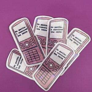 May include: A collection of pink and white stickers shaped like retro cell phones. Each sticker has the text "Be gentle - I survived the 1900s" in black. The stickers are arranged on a purple background.