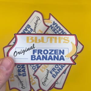 Könnte beinhalten: Mehrere Aufkleber auf gelbem Hintergrund. Die Aufkleber zeigen den Text "BLUTH'S Original FROZEN BANANA" in Blau und Gelb, mit einer gelben Bananengrafik. Die Aufkleber haben einen weißen und roten Rand.
