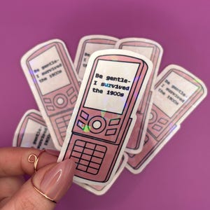 May include: Pink stickers featuring a retro flip phone design with the text "Be gentle - I survived the 1900s." The stickers have a holographic effect and are arranged on a purple background.