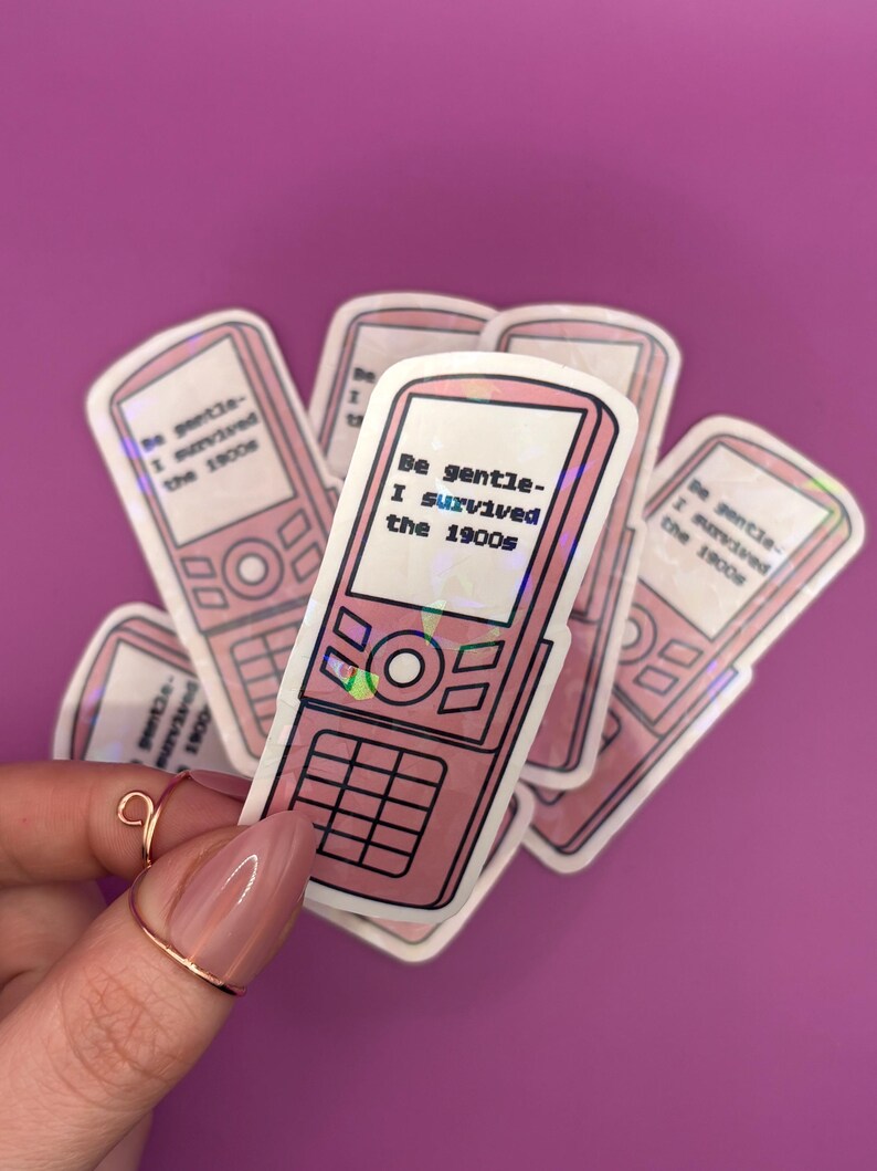 May include: Pink stickers featuring a retro flip phone design with the text "Be gentle - I survived the 1900s." The stickers have a holographic effect and are arranged on a purple background.