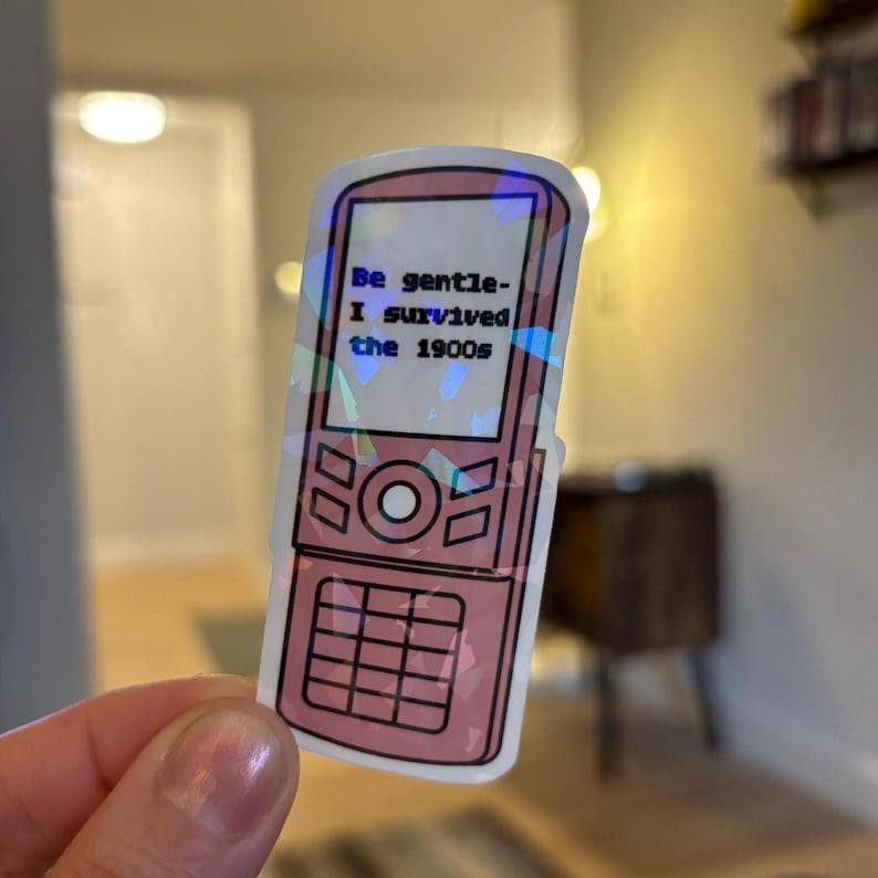 May include: A pink sticker in the shape of a flip phone with the text "Be gentle- I survived the 1900s." The sticker has a holographic effect and black outlines. The phone design includes buttons and a circular center button.