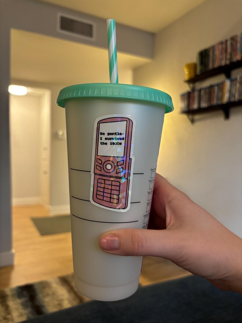 May include: A reusable plastic tumbler with a green lid and straw. A sticker of a pink flip phone with the text "Be gentle- I survived the 1900s" is affixed to the cup. The cup is held by a person.