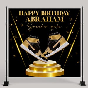 May include: A black and gold banner with the text "HAPPY BIRTHDAY ABRAHAM Sneaker gala." Two black and gold high-top sneakers are displayed on a golden tiered platform. The background is black with gold star and light accents.