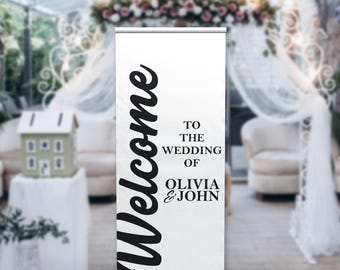 May include: A white custom retractable banner with black text that reads "Welcome" and "To the Wedding of Olivia & John." The banner is displayed at a wedding venue with floral decorations and seating.
