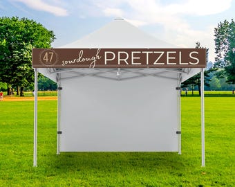 May include: A white pop-up canopy tent featuring a brown valance with the text 47 SOURDOUGH PRETZELS printed in white. The tent has a metal frame and a solid white back wall, standing on a green lawn under a bright blue sky with scattered clouds.