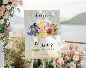 May include: A baby shower sign featuring Winnie the Pooh and friends. The sign reads "Welcome to Marie's Baby Shower" with the date 10-10-25. The sign is on an easel, surrounded by flowers and greenery.