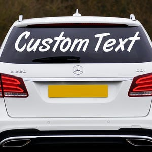 May include: White car with a rear window decal that reads "Custom Text" in a cursive font. The car has red taillights and a yellow license plate. The car is a Mercedes-Benz.