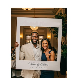 May include: A white photo booth frame with the text "Name & Name" and "xx.xx.20xx" at the bottom. A smiling couple poses inside the frame. The background features a well-lit room with chandeliers.