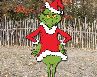 May include: A green Grinch character cutout wearing a red Santa suit with white trim and a matching hat. The Grinch has yellow eyes and is standing in front of a wooden fence and fall foliage.