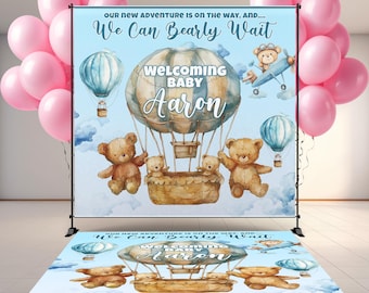 May include: A custom backdrop and floor decal set for a baby shower. The backdrop is light blue with hot air balloons, teddy bears, and the text "Welcoming Baby Aaron." Pink balloons are in the background.