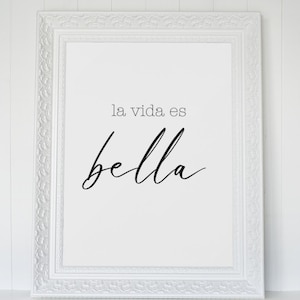 May include: White framed print with the text "la vida es bella" in black cursive script.