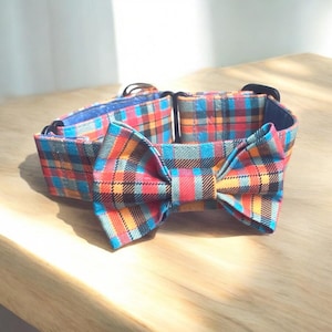 May include: A colorful plaid dog collar with a matching bow tie. The collar features a pattern of red, orange, blue, and black squares. The bow tie is centered on the collar. The collar is made of fabric and has a blue interior.
