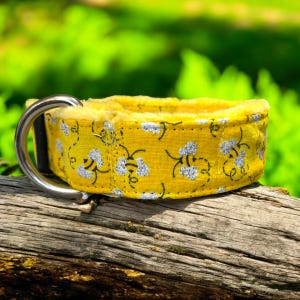 May include: Yellow dog collar with a bee pattern. The collar features a silver metal D-ring and a soft, light yellow interior. The bee pattern has white and black bees on a yellow background.