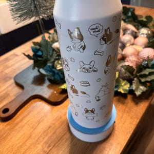 Pu&ograve; includere: Una borraccia isolante bianca con un motivo dorato di cani e ossa. La bottiglia ha un coperchio bianco e poggia su una base bianca e blu. Le parole "Woof! Woof!" sono stampate sulla bottiglia.