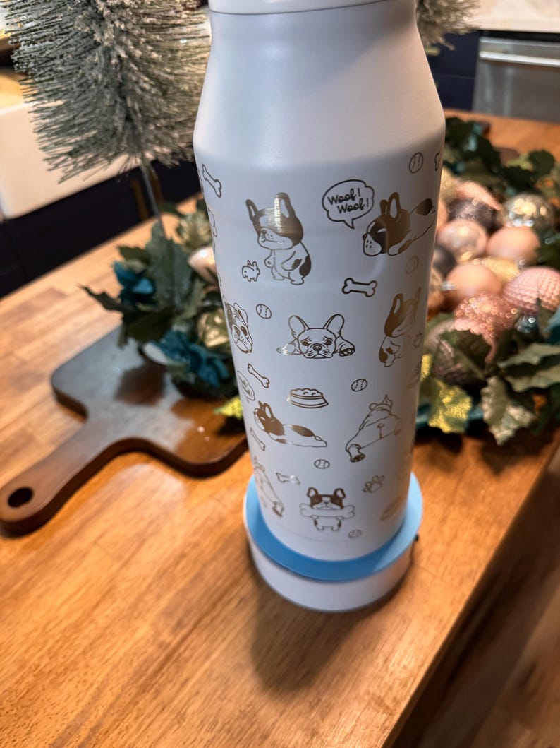 Pu&ograve; includere: Una borraccia isolante bianca con un motivo dorato di cani e ossa. La bottiglia ha un coperchio bianco e poggia su una base bianca e blu. Le parole "Woof! Woof!" sono stampate sulla bottiglia.