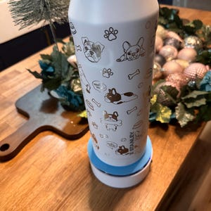 Pu&ograve; includere: Una borraccia Stanley bianca con base azzurra, decorata con illustrazioni dorate di cani, impronte di zampe e ossa. La borraccia cilindrica poggia su una base di ricarica bianca e blu. La parola "Stanley" &egrave; visibile sulla borraccia.