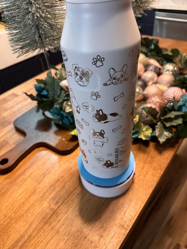 Pu&ograve; includere: Una borraccia Stanley bianca con base azzurra, decorata con illustrazioni dorate di cani, impronte di zampe e ossa. La borraccia cilindrica poggia su una base di ricarica bianca e blu. La parola "Stanley" &egrave; visibile sulla borraccia.