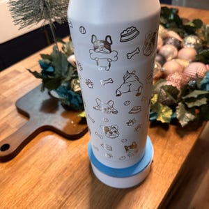 Pu&ograve; includere: Una borraccia isolante bianca con un motivo dorato a tema cani. Il motivo include facce di cani, ossa, impronte di zampe e ciotole per cibo. La bottiglia &egrave; su una base bianca e blu e una superficie di legno.