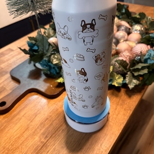 Pu&ograve; includere: Una bottiglia d'acqua isolante bianca decorata con illustrazioni dorate di cani, tra cui bulldog francesi e ossa. La bottiglia &egrave; posta su una base bianca e blu. Ideale per gli amanti dei cani.