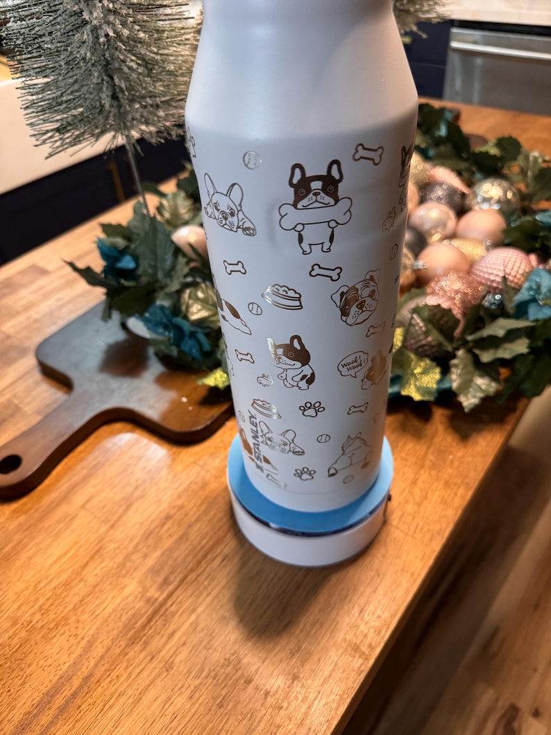 Pu&ograve; includere: Una bottiglia d'acqua isolante bianca decorata con illustrazioni dorate di cani, tra cui bulldog francesi e ossa. La bottiglia &egrave; posta su una base bianca e blu. Ideale per gli amanti dei cani.