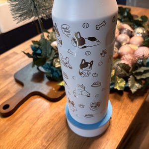 Pu&ograve; includere: Una bottiglia d'acqua isolante bianca con illustrazioni dorate di bulldog francesi, ossa e impronte di zampe. La bottiglia ha una base azzurra e un coperchio argentato. Le parole "Woof! Woof!" sono stampate.