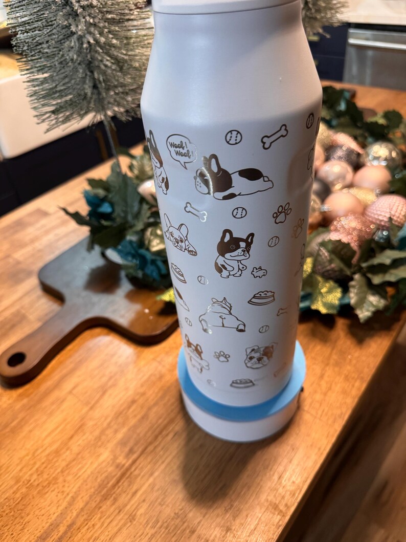 Pu&ograve; includere: Una bottiglia d'acqua isolante bianca con illustrazioni dorate di bulldog francesi, ossa e impronte di zampe. La bottiglia ha una base azzurra e un coperchio argentato. Le parole "Woof! Woof!" sono stampate.