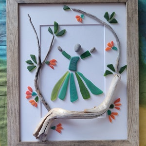 May include: A framed piece of art made with sea glass and driftwood. The art depicts a stylized figure in a green dress with orange flowers and green leaves around it.