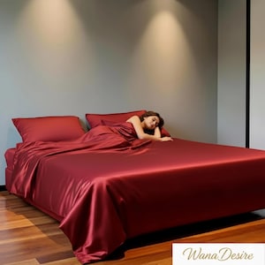 Puede incluir: Una cama con un juego de sábanas de satén rojo intenso. El juego incluye una sábana bajera, una sábana encimera y fundas de almohada. Una persona está acostada en la cama, cubierta por la sábana. La marca "Wana Desire" es visible en la esquina inferior derecha.