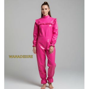 Könnte beinhalten: Ein leuchtend pinkfarbener Overall mit hohem Rüschenkragen und langen Ärmeln. Das glänzende, einteilige Outfit hat ein eng anliegendes Oberteil und eine locker sitzende Hose. Der Text "WANADESIRE" ist unten links sichtbar.