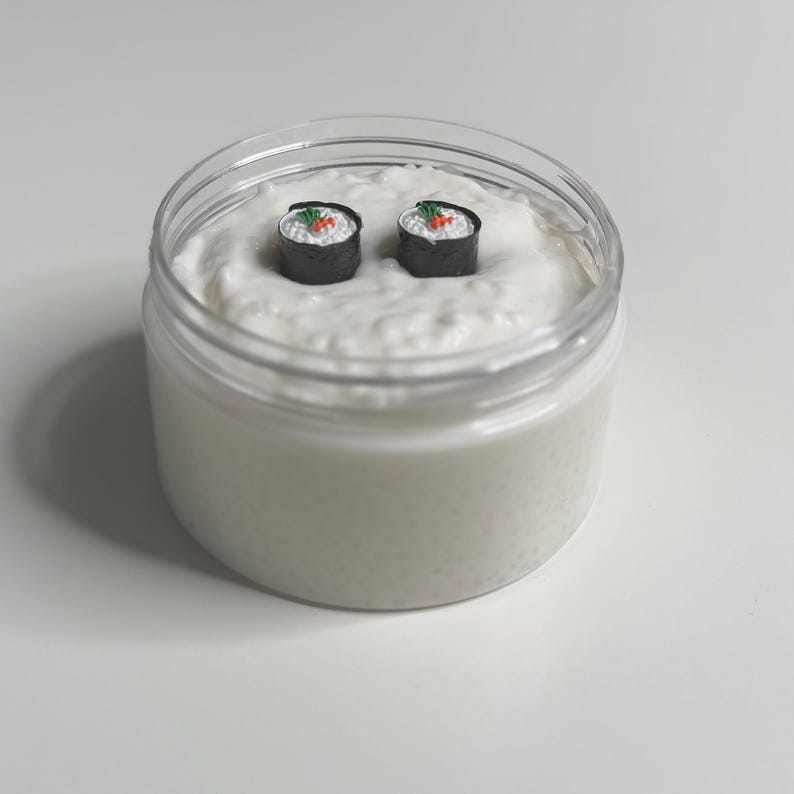 Pode incluir: Uma subst&acirc;ncia cremosa branca em um frasco transparente com dois pequenos rolinhos de sushi no topo.