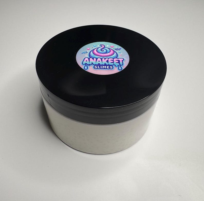 Pode incluir: Um recipiente de slime branco com uma tampa preta. A tampa tem um adesivo colorido com o texto "Anakeet Slimes".