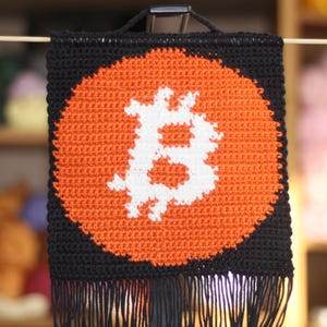 May include: A black crochet wall hanging featuring an orange circle with a white Bitcoin symbol. The design has black fringe at the bottom and a wooden dowel for hanging. The background is blurred, with shelves of yarn visible.