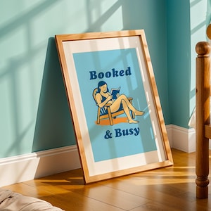 May include: A framed art print with a light blue background and the words "Booked & Busy" in navy blue. The print features a woman reading in a chair. The frame is made of light-colored wood.