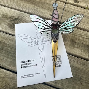 Może przedstawiać: Witrołazowy sztylet cykady z iryzującymi skrzydłami, różowym i fioletowym uchwytem oraz złotym ostrzem. Sztylet jest zawieszony na łańcuchu. Wzór sztyletu znajduje się na białej kartce z napisem "@KOZLOOPERGLASS Cicada Dagger Stained Glass Pattern".