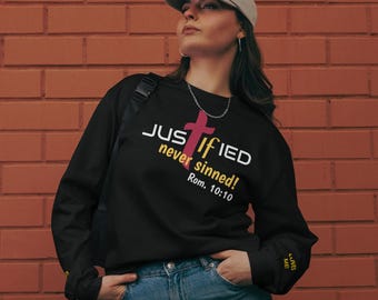 May include: Black sweatshirt with the text "JUSTIFIED never sinned!" and a pink cross graphic. The text "Rom. 10:10" is below the graphic. The sweatshirt has long sleeves and is worn with blue jeans and a gray cap.