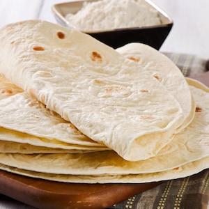 May include: A stack of round, thin, off-white flour tortillas with light brown toasted spots. The tortillas are layered on top of each other, showing a soft, pliable texture. Each tortilla has a slightly irregular circular shape with visible surface bubbles.