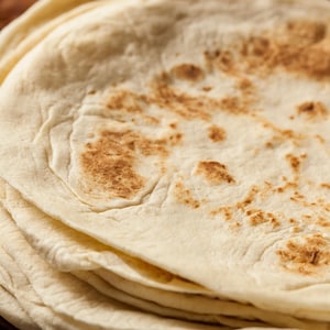 May include: A stack of fresh, homemade tortillas. The tortillas are a light beige color with areas of golden-brown browning. The edges of the tortillas are slightly uneven, and the texture appears soft and slightly bubbly.