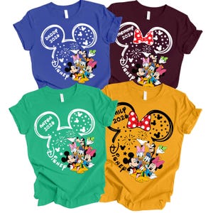 Puede incluir: Cuatro camisetas coloridas con tem&aacute;tica de Disney. Cada camiseta presenta un dise&ntilde;o de cabeza de Mickey Mouse con texto y personajes de Disney. Los colores incluyen azul, granate, verde y amarillo. El texto incluye "Daddy 2026", "Mommy 2026", "Anton 2026" y "Family 2026".
