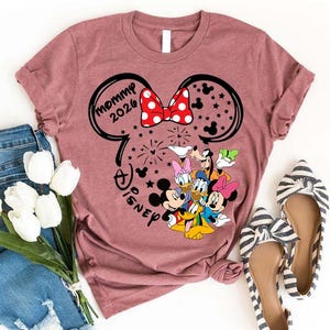 Puede incluir: Una camiseta de color malva con un dise&ntilde;o inspirado en Minnie Mouse, con el texto "Mommy 2026" y el logotipo de Disney. El dise&ntilde;o incluye a Mickey y Minnie Mouse, Donald y Daisy Duck, y Goofy. La camiseta se combina con vaqueros azules y bailarinas a rayas.