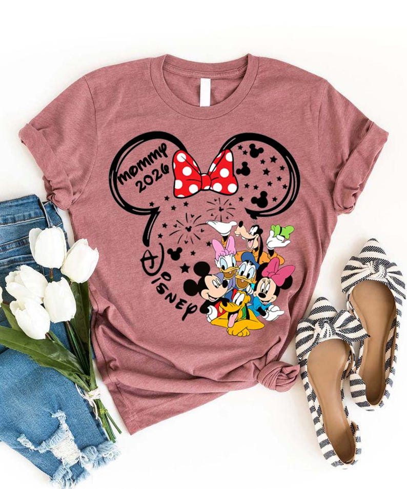 Puede incluir: Una camiseta de color malva con un dise&ntilde;o inspirado en Minnie Mouse, con el texto "Mommy 2026" y el logotipo de Disney. El dise&ntilde;o incluye a Mickey y Minnie Mouse, Donald y Daisy Duck, y Goofy. La camiseta se combina con vaqueros azules y bailarinas a rayas.