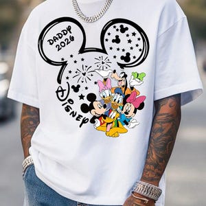 Puede incluir: Camiseta blanca con un dise&ntilde;o negro de la cabeza de Mickey Mouse, el texto "DADDY 2026" y "Disney". El dise&ntilde;o incluye los personajes de dibujos animados Mickey, Minnie, Donald, Daisy, Goofy y Pluto. La camiseta tiene mangas cortas.