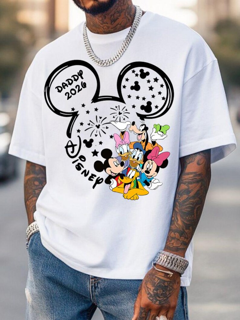Puede incluir: Camiseta blanca con un dise&ntilde;o negro de la cabeza de Mickey Mouse, el texto "DADDY 2026" y "Disney". El dise&ntilde;o incluye los personajes de dibujos animados Mickey, Minnie, Donald, Daisy, Goofy y Pluto. La camiseta tiene mangas cortas.