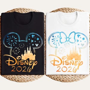 May include: A black t-shirt and a white t-shirt, each featuring a graphic of a castle silhouette inside a Mickey Mouse ear outline with fireworks, stars, and the text Disney 2026. The white shirt includes a bow on the Mickey ear outline.
